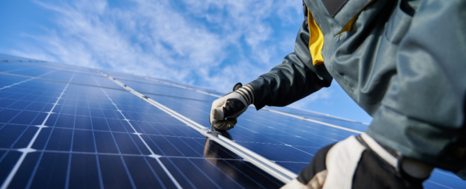 A person in protective gear and gloves is inspecting or installing a solar panel under a clear blue sky.