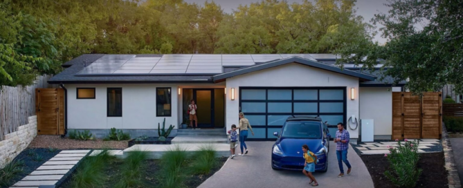 This image depicts a modern, single-story home with solar panels installed on the roof. A family is seen in the driveway: a woman standing at the front door, a man standing near a blue Tesla Model Y parked in the driveway, and two children playing nearby. The home features a sleek design with a mix of white and dark exterior finishes, a glass garage door, and a well-manicured front yard with plants and trees. Additionally, a Tesla Powerwall is mounted on the exterior wall near the garage. The setting appears to be a suburban neighborhood, emphasizing a clean and eco-friendly lifestyle.